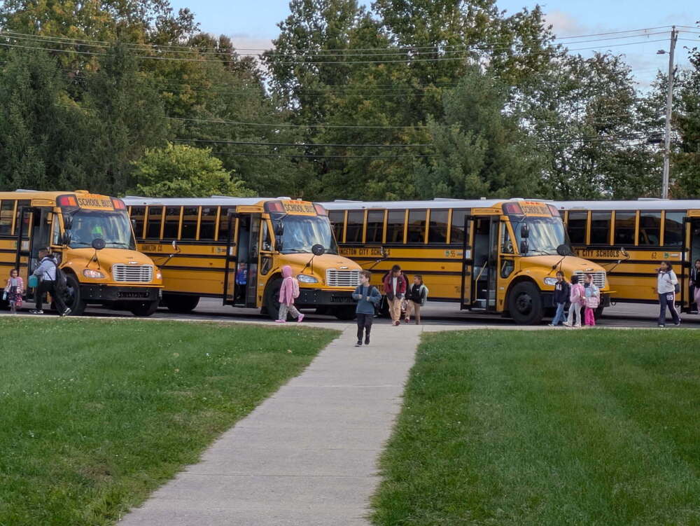 Students walk away from three parked yellow school buses on a paved path, with adults nearby and trees in the background.