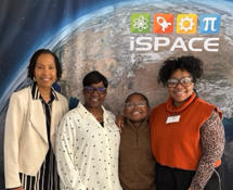 Four people stand smiling in front of an iSPACE banner showing Earth from space: teacher Donna Powell in a cream jacket, two additional women, and a Woodlawn student in a brown sweater and glasses.
