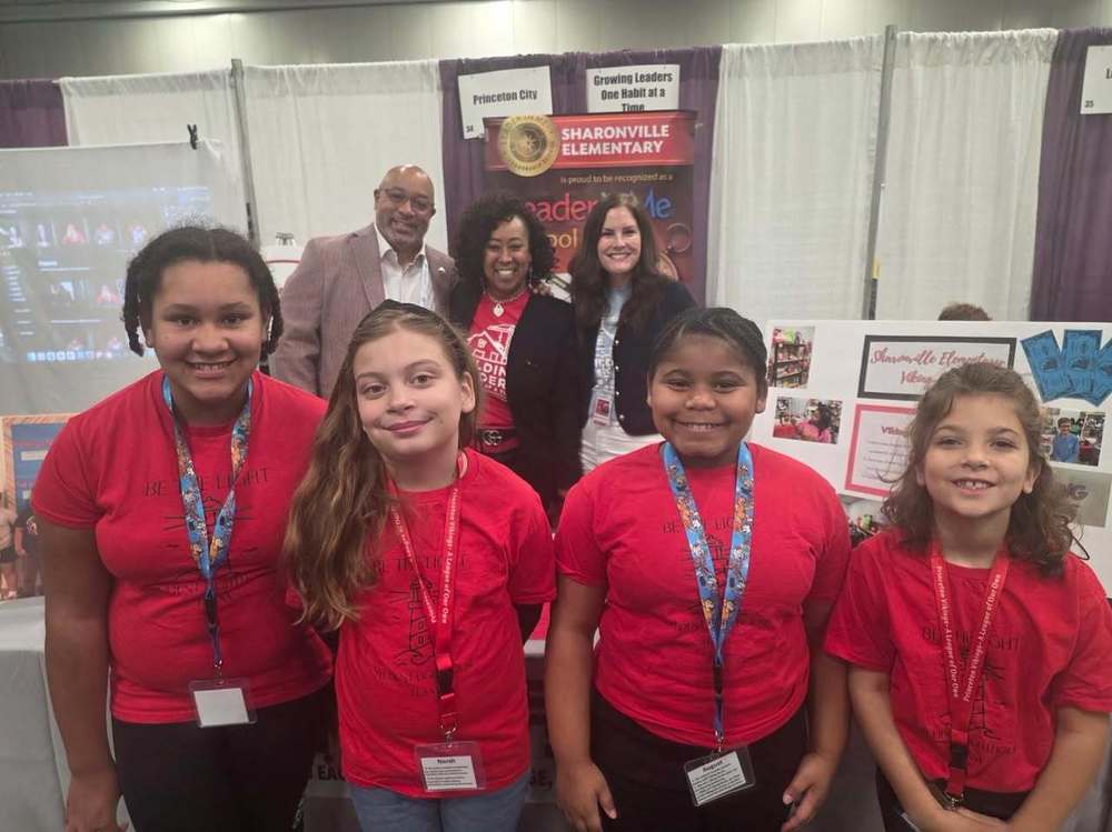 Four Sharonville elementary students wearing matching red shirts stand and smile for the camera at a school event booth. Behind them, three adults also smile, standing in front of a display for Sharonville Elementary with posters and banners highlighting school programs. The booth features colorful visuals, student projects, and informational materials. The setting appears to be an indoor conference.