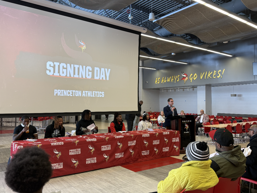 Signing Day athletes  sitting at table with Princeton Viking logos and Mr. John Wilhelm, address the seniors