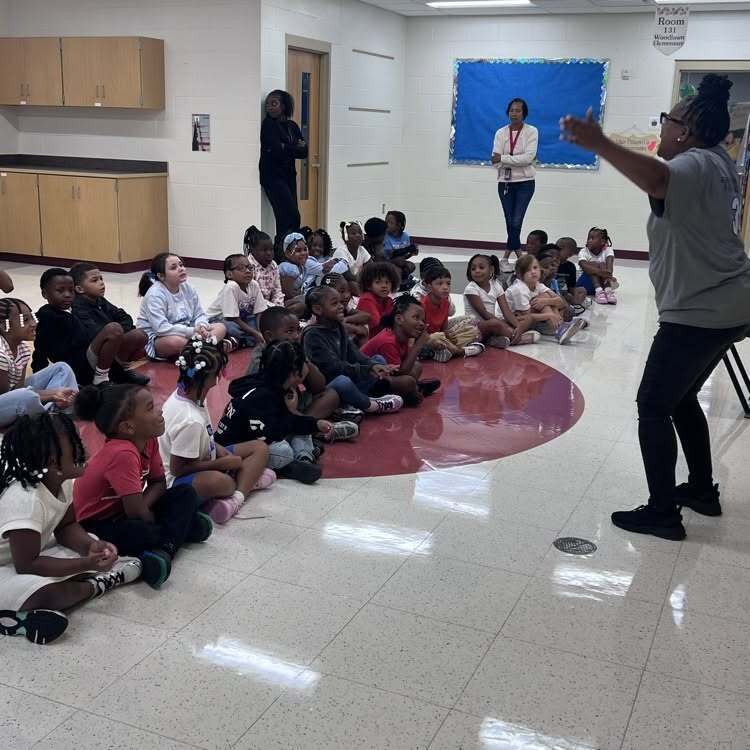 Woodlawn Principal Sherry Thompson gives animated talk to students on the first day of school. More than a dozen students are sitting on the ground with crossed legs in front of Principal Thompson.