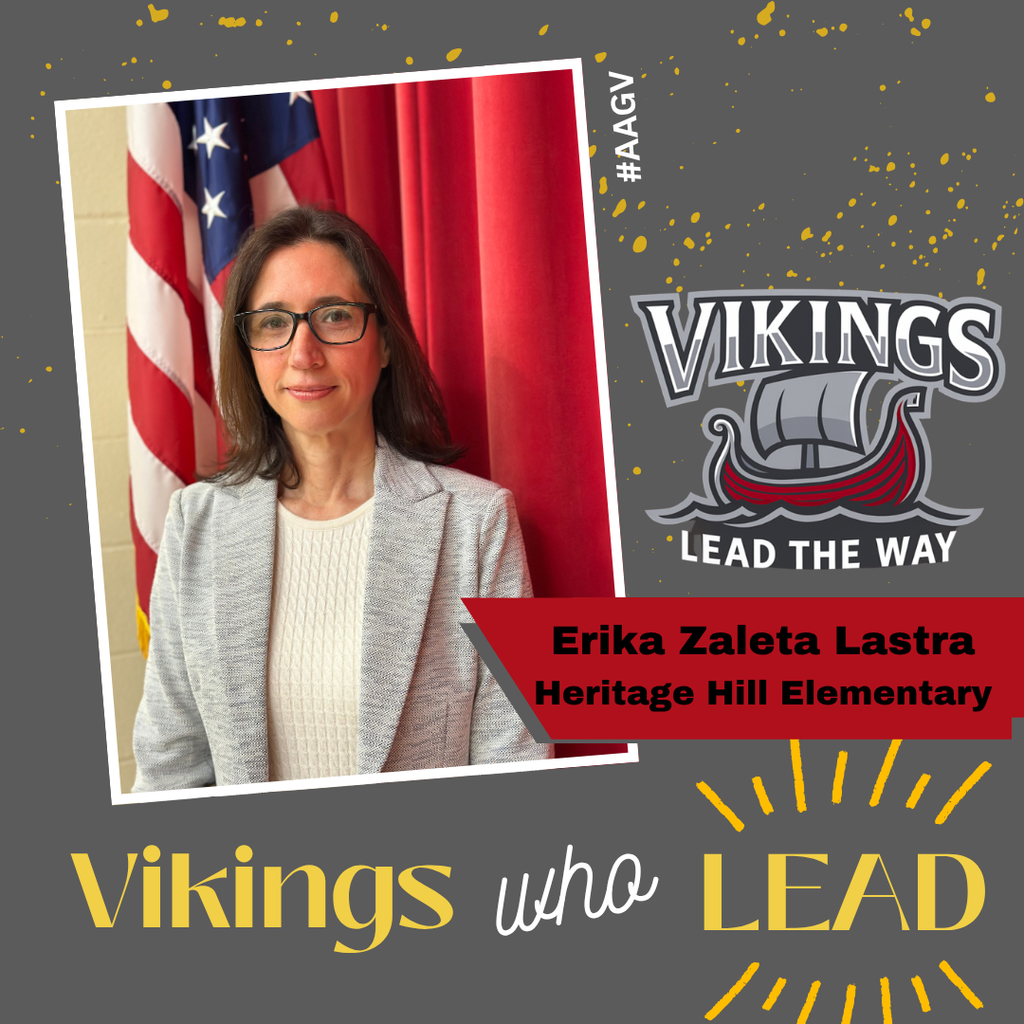 Professional photograph of Erika Zaleta Lastra from Heritage Hill Elementary wearing glasses, a cream-colored cable knit sweater, and a light gray blazer, standing in front of an American flag and red curtain backdrop. The image is displayed in a tilted white frame against a gray background decorated with gold confetti. The Vikings logo featuring a viking ship with 'Lead The Way' motto appears in the top right. Text at bottom reads 'Vikings who LEAD' with 'Vikings' and 'LEAD' in gold and 'who' in white script. A red banner identifies 'Erika Zaleta Lastra, Heritage Hill Elementary.' Hashtag #AAGY appears vertically along the right edge.