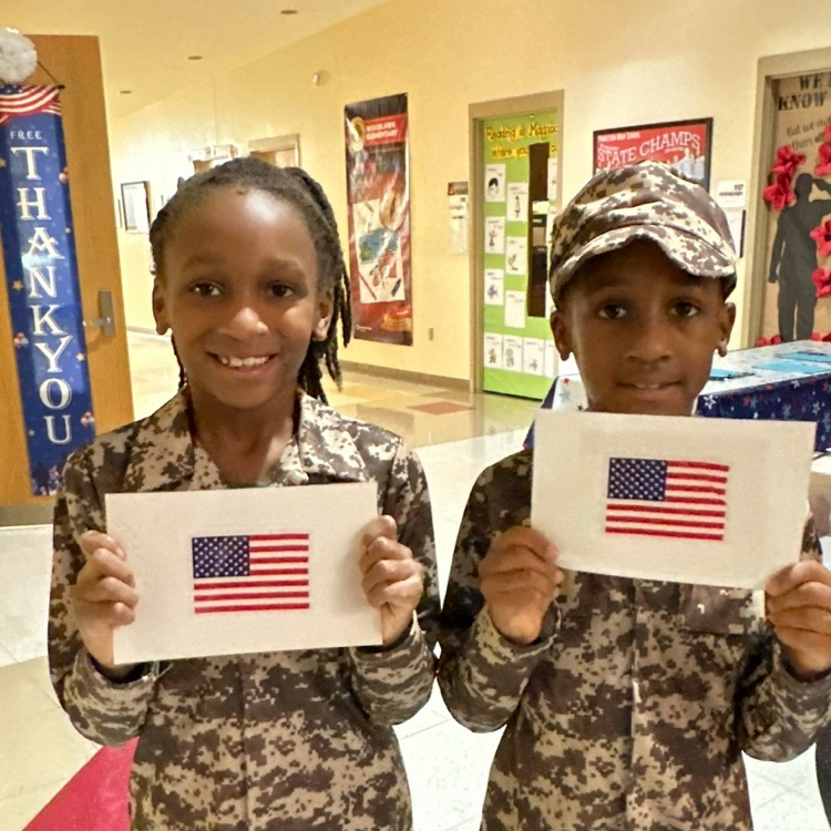 Two military family students dressed in camo holding print outs of an American flag. Three doors behind are decorated to celebrate Veterans Day. one says Thank You. 