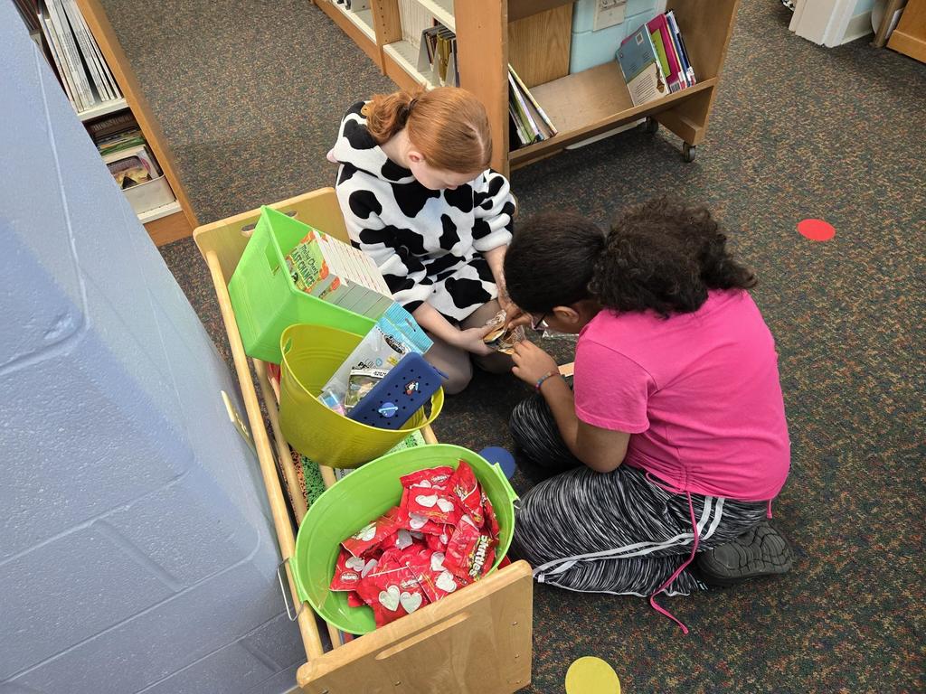 #EV Two children sitting on a library floor looking at an object beside a low bookshelf with bins of books and snacks.