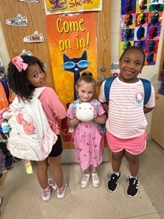 Two young children stand side by side in a classroom, smiling at the camera. They are wearing matching floral tops; one has shorts and sneakers, and the other has a pink skirt and sparkly shoes. A colorful painted door with a cat design is behind them.