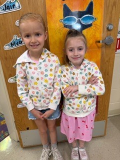 Two young children stand side by side in a classroom, smiling at the camera. They are wearing matching floral tops; one has shorts and sneakers, and the other has a pink skirt and sparkly shoes. A colorful painted door with a cat design is behind them.