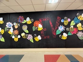 A school hallway wall display featuring a black background decorated with colorful paper flowers, yellow-and-black bees, green leaves, and large red letters spelling “LOVE” across the center. The floor has multicolored geometric tiles, creating a bright and cheerful atmosphere. 