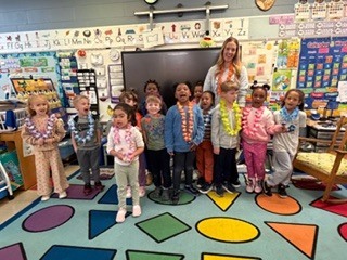 Group of young children standing in a classroom, many wearing colorful leis, with a teacher smiling behind them; bright alphabet posters and a multicolored shape rug are visible.