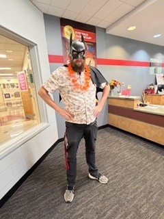 Adult standing in an office space wearing a black Batman-style mask and an orange lei, hands on hips, with a service counter and hallway visible in the background