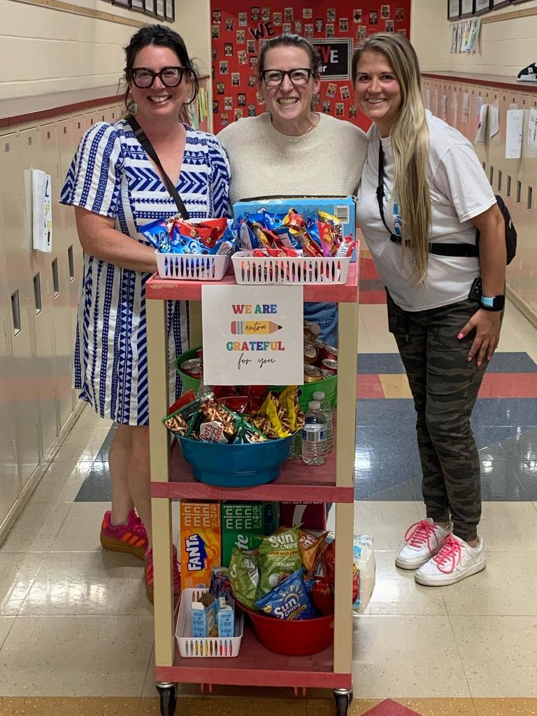 Three women smiling in a school hallway stand behind a rolling cart filled with snacks and drinks. A sign on the cart reads, “We are extra grateful for you.” Lockers line the hallway, and a red bulletin board with photos is visible in the background.