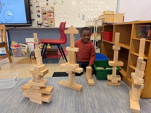 A young student sits on a classroom rug surrounded by several tall wooden block towers he has built. The classroom includes shelves, bins, a red chair, and a screen displaying a clock activity in the background. The student appears focused and engaged in building and balancing the structures.