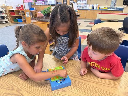 Two young children play together at a red sensory table filled with sand and small ocean-themed toys, including turtles, shells, and sea animals. The children are focused on scooping and exploring the sand while standing in a classroom environment.