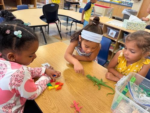 Two young children play together at a red sensory table filled with sand and small ocean-themed toys, including turtles, shells, and sea animals. The children are focused on scooping and exploring the sand while standing in a classroom environment.