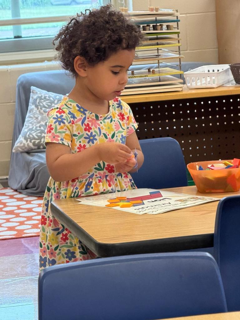 Two young children play together at a red sensory table filled with sand and small ocean-themed toys, including turtles, shells, and sea animals. The children are focused on scooping and exploring the sand while standing in a classroom environment.