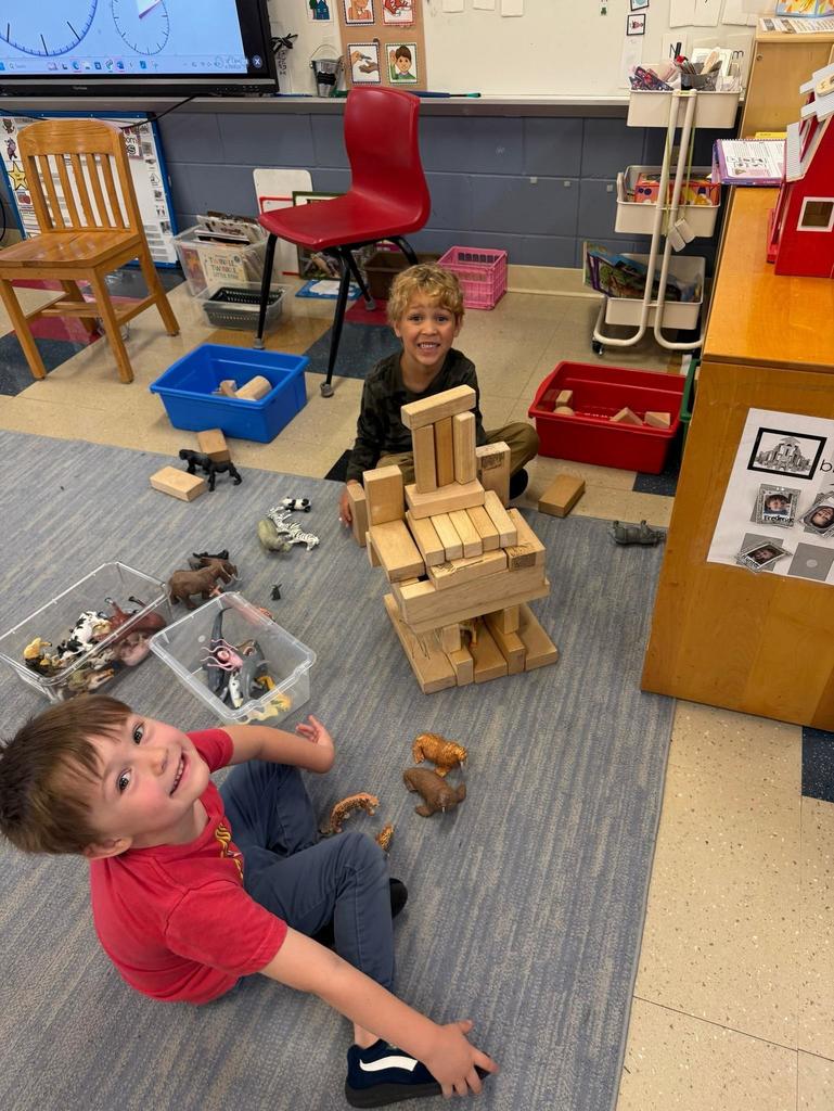 A young student sits on a classroom rug surrounded by several tall wooden block towers he has built. The classroom includes shelves, bins, a red chair, and a screen displaying a clock activity in the background. The student appears focused and engaged in building and balancing the structures.