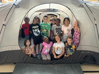 A group of elementary school students sits and kneels in a school room in front of a large gray camping tent. The students smile and pose together, with colorful artwork displayed on the walls behind them and sunlight coming through high windows, creating a bright and cheerful indoor setting.