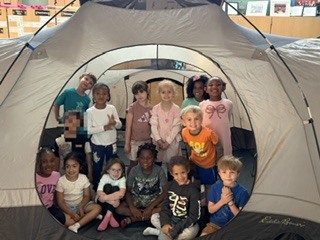 A group of elementary school students sits and kneels in a school room in front of a large gray camping tent. The students smile and pose together, with colorful artwork displayed on the walls behind them and sunlight coming through high windows, creating a bright and cheerful indoor setting.