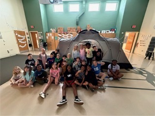 A group of elementary school students sits and kneels in a school room in front of a large gray camping tent. The students smile and pose together, with colorful artwork displayed on the walls behind them and sunlight coming through high windows, creating a bright and cheerful indoor setting.