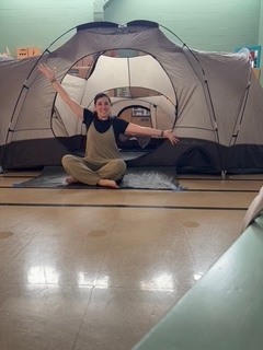 A group of elementary school students sits and kneels in a school room in front of a large gray camping tent. The students smile and pose together, with colorful artwork displayed on the walls behind them and sunlight coming through high windows, creating a bright and cheerful indoor setting.