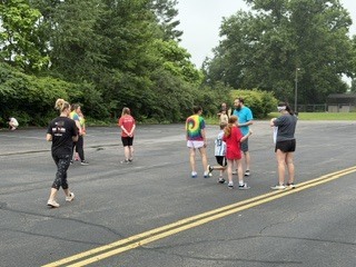 A group of children and adults stand together on a paved outdoor area surrounded by trees during an overcast day. Several children wear colorful shirts while adults appear to supervise or organize an activity. Yellow road lines run through the center of the pavement, and the setting resembles a school or park recreation area.