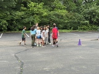 A group of children and adults stand together on a paved outdoor area surrounded by trees during an overcast day. Several children wear colorful shirts while adults appear to supervise or organize an activity. Yellow road lines run through the center of the pavement, and the setting resembles a school or park recreation area.