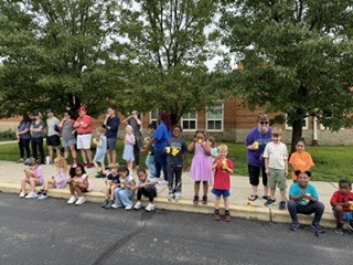 A group of children and adults stand together on a paved outdoor area surrounded by trees during an overcast day. Several children wear colorful shirts while adults appear to supervise or organize an activity. Yellow road lines run through the center of the pavement, and the setting resembles a school or park recreation area.