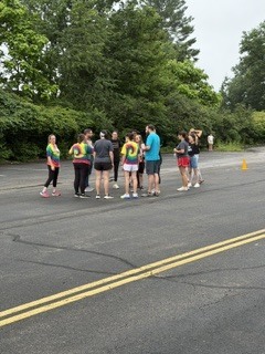 A group of children and adults stand together on a paved outdoor area surrounded by trees during an overcast day. Several children wear colorful shirts while adults appear to supervise or organize an activity. Yellow road lines run through the center of the pavement, and the setting resembles a school or park recreation area.