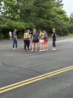 A group of children and adults stand together on a paved outdoor area surrounded by trees during an overcast day. Several children wear colorful shirts while adults appear to supervise or organize an activity. Yellow road lines run through the center of the pavement, and the setting resembles a school or park recreation area.