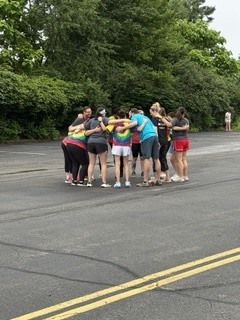 A group of children and adults stand together on a paved outdoor area surrounded by trees during an overcast day. Several children wear colorful shirts while adults appear to supervise or organize an activity. Yellow road lines run through the center of the pavement, and the setting resembles a school or park recreation area.
