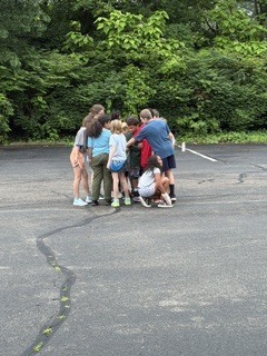 A group of children and adults stand together on a paved outdoor area surrounded by trees during an overcast day. Several children wear colorful shirts while adults appear to supervise or organize an activity. Yellow road lines run through the center of the pavement, and the setting resembles a school or park recreation area.