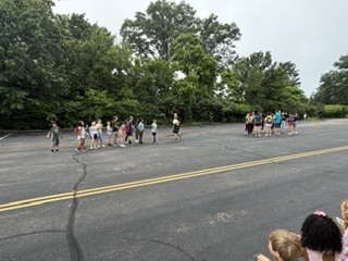 A group of children and adults stand together on a paved outdoor area surrounded by trees during an overcast day. Several children wear colorful shirts while adults appear to supervise or organize an activity. Yellow road lines run through the center of the pavement, and the setting resembles a school or park recreation area.