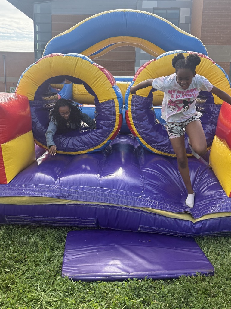 A child leaps out of a colorful inflatable obstacle course while an adult emerges from a tunnel beside her, both smiling and having fun outdoors on a grassy area near a brick school building.