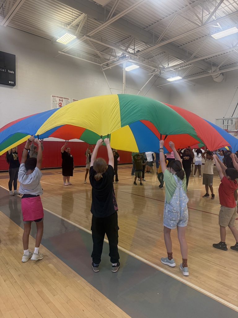 A large group of children and adults stand in a circle inside a gymnasium, lifting a large colorful parachute high above their heads together.