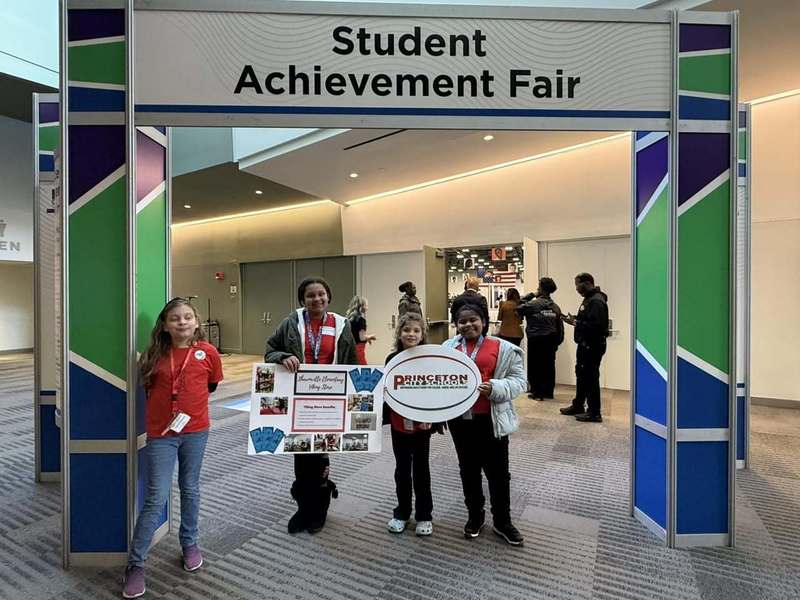 Four elementary students stand under a large “Student Achievement Fair” sign in a convention center hallway.