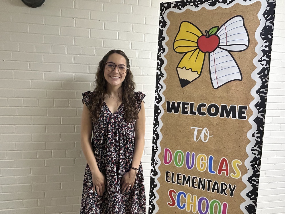 photo of Mrs. Anna Aughenbaugh-Weber standing next to a sign reading "Welcom to Douglas Elementary School"