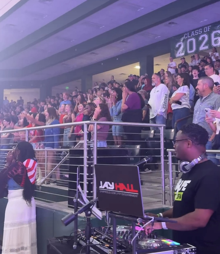 Student in gym arena stands standing and yelling. A DJ stands at his station watching on