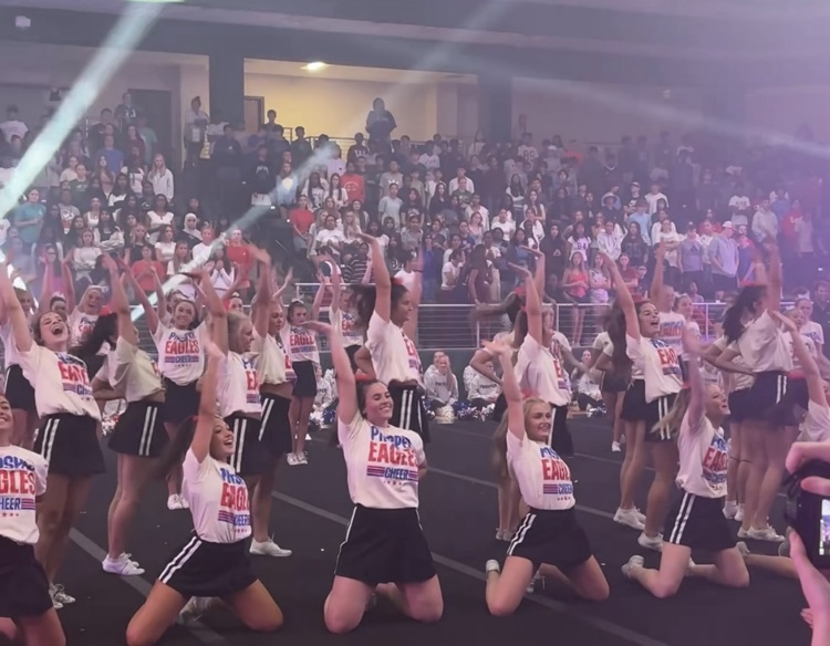 Cheerleaders pose during a dance with their right hands raised and smiling expressions. The stands are full behind them