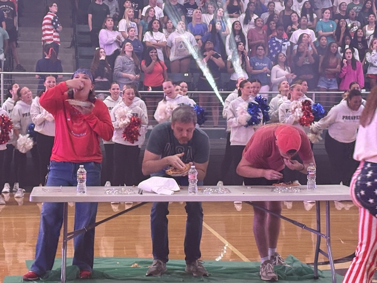 Three people participate in a pie eating contest. One is standing, two are bent over a table, all eating pie with their hands