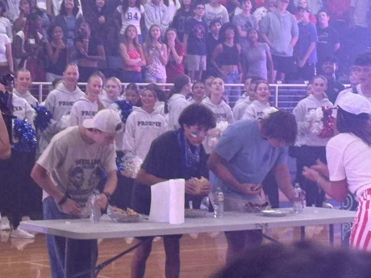 Three people participate in a pie eating contest. One is standing, two are bent over a table, all eating pie with their hands
