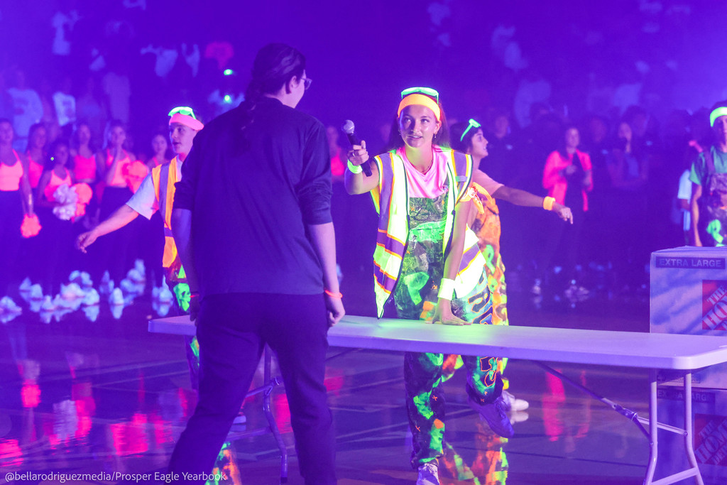 Students in neon vests and overalls host a game on the gym floor under glowing lights.