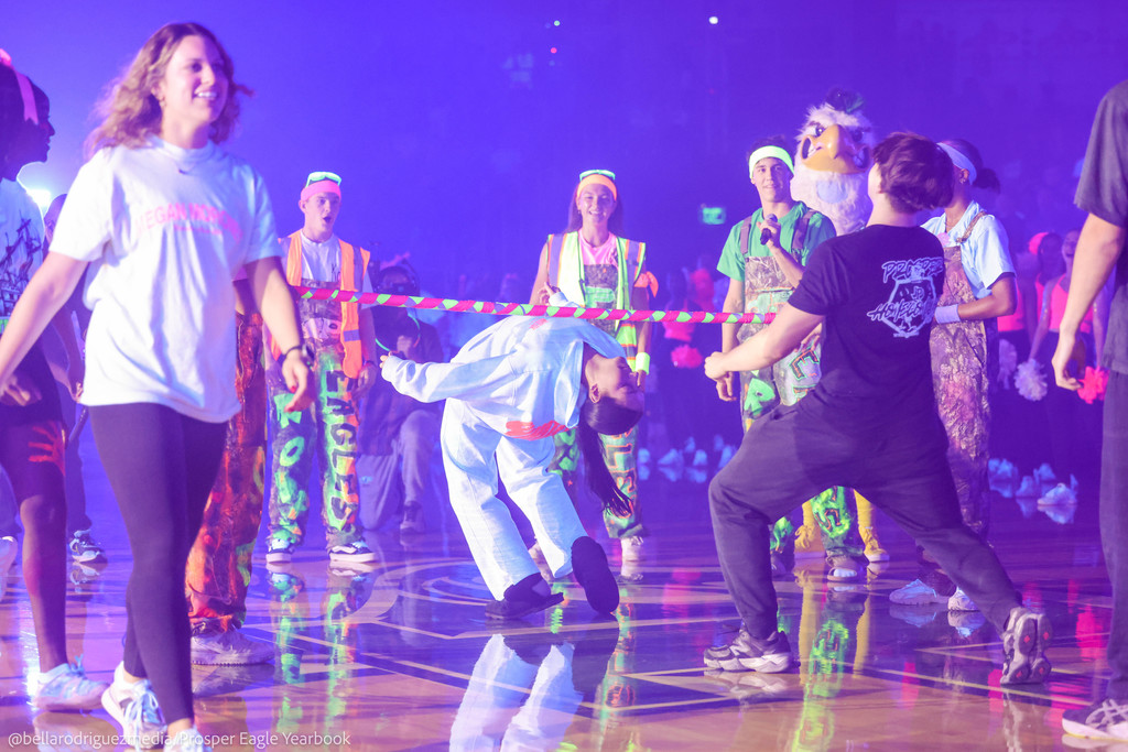 Students participate in a neon-themed pep rally limbo contest under blacklights, surrounded by glowing outfits and decorations.