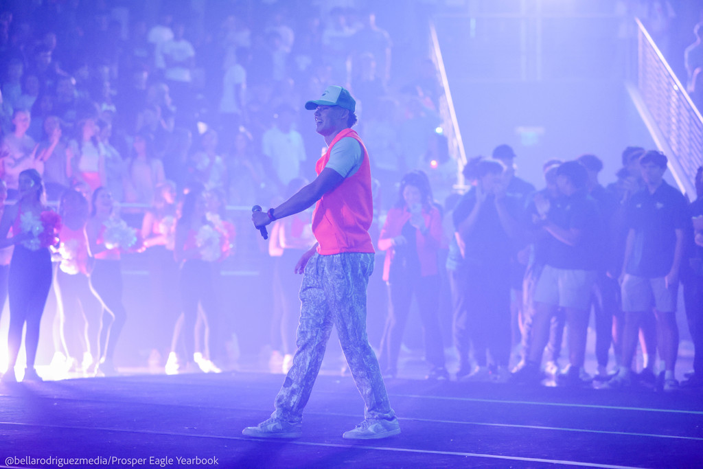 Student MC in neon gear speaks into a microphone, with the crowd and cheer team cheering in the background.