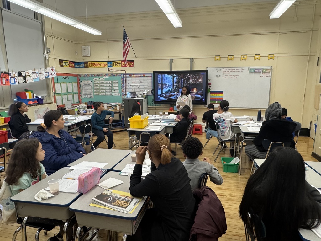 Student reads from a paper, at the front of a classroom, while standing in front of an image of a fireplace in a cozy living room during wintertime, while other students and parents look on and listen