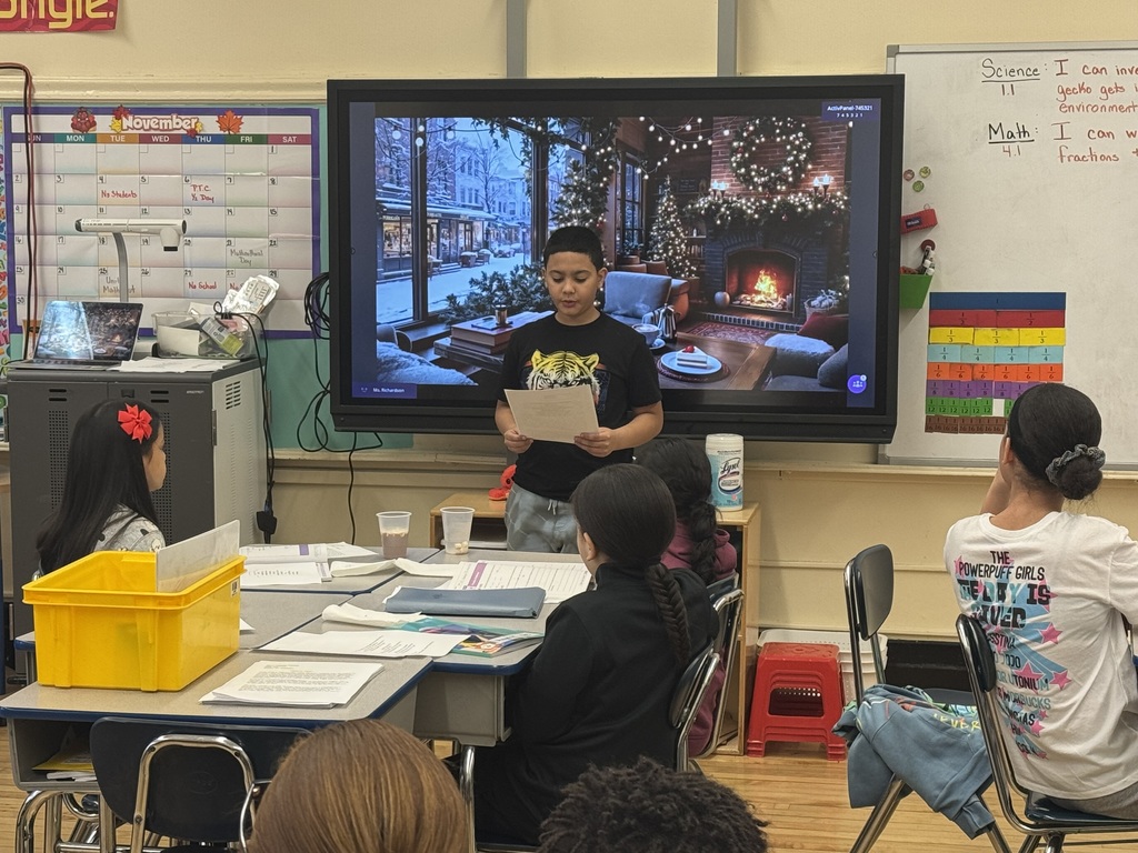 Student reads from a paper, at the front of a classroom, while standing in front of an image of a fireplace in a cozy living room during wintertime, while other students look on and listen