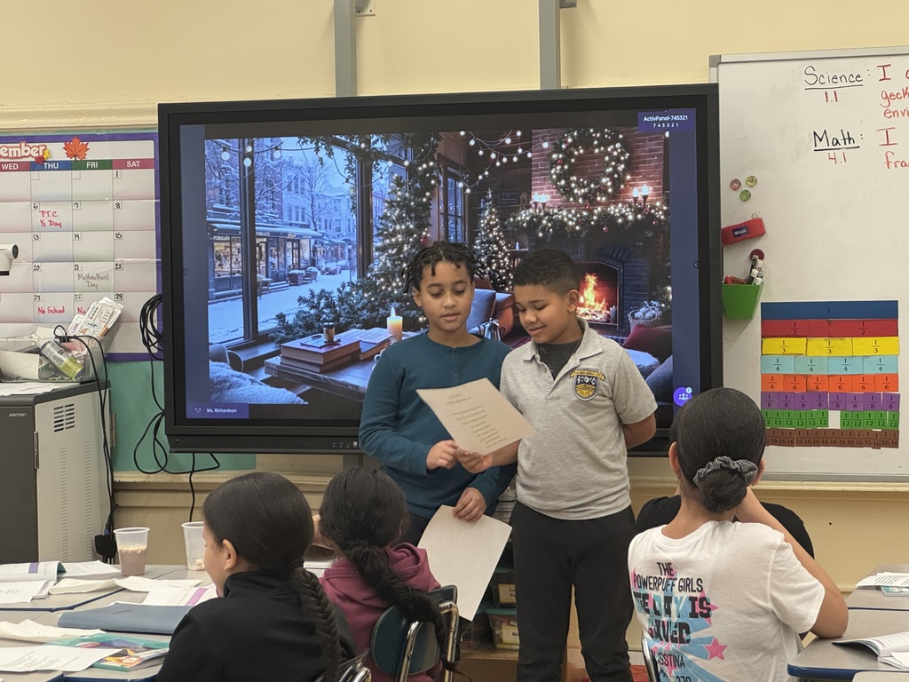 Two students reads from a paper, at the front of a classroom, while standing in front of an image of a fireplace in a cozy living room during wintertime, while other students look on and listen