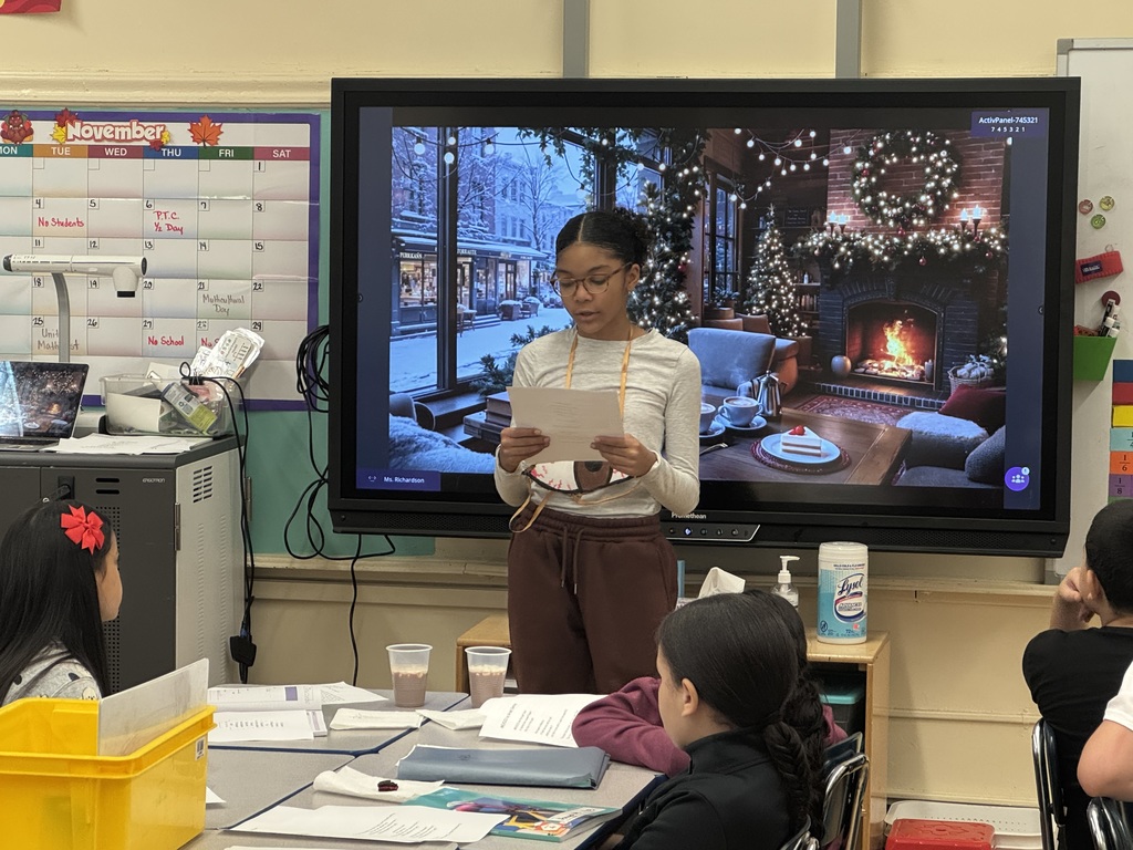 Student reads from a paper, at the front of a classroom, while standing in front of an image of a fireplace in a cozy living room during wintertime, while other students look on and listen
