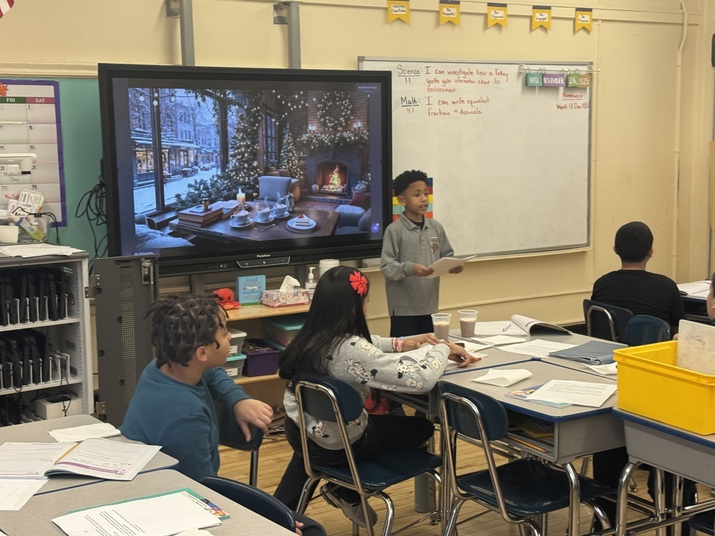 Student reads from a paper, at the front of a classroom, while standing in front of an image of a fireplace in a cozy living room during wintertime, while other students look on and listen