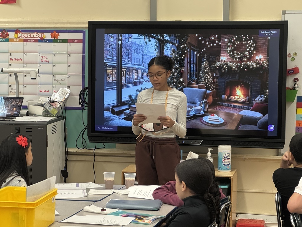 Student reads from a paper, at the front of a classroom, while standing in front of an image of a fireplace in a cozy living room during wintertime, while other students look on and listen