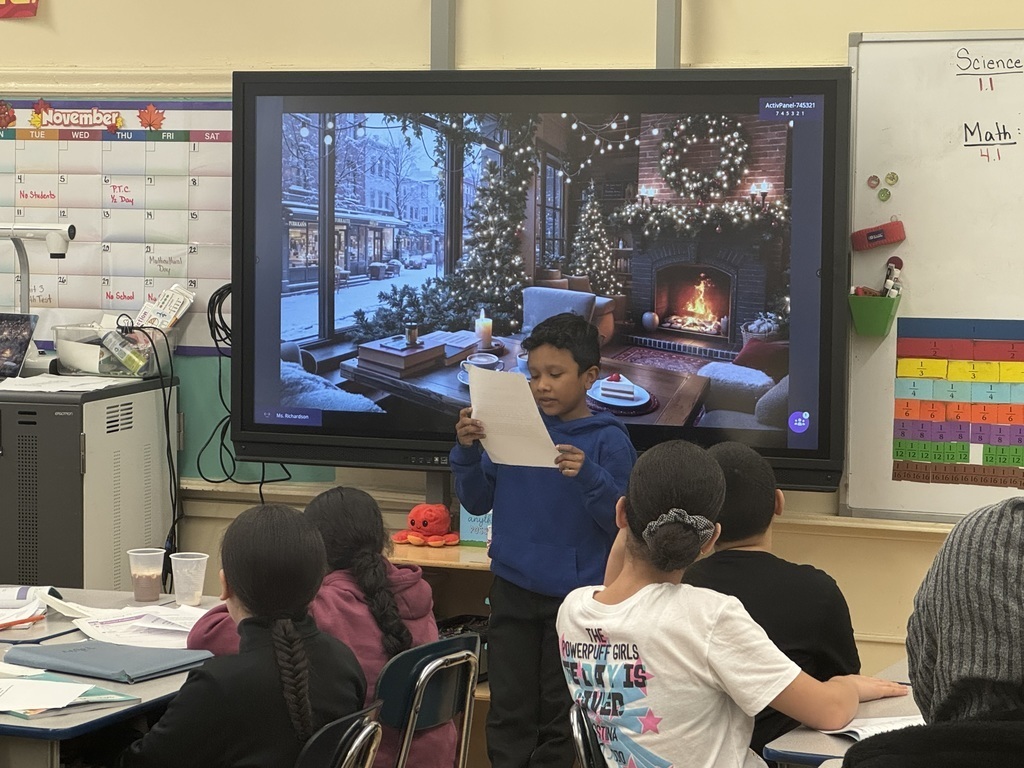 Student reads from a paper, at the front of a classroom, while standing in front of an image of a fireplace in a cozy living room during wintertime, while other students look on and listen