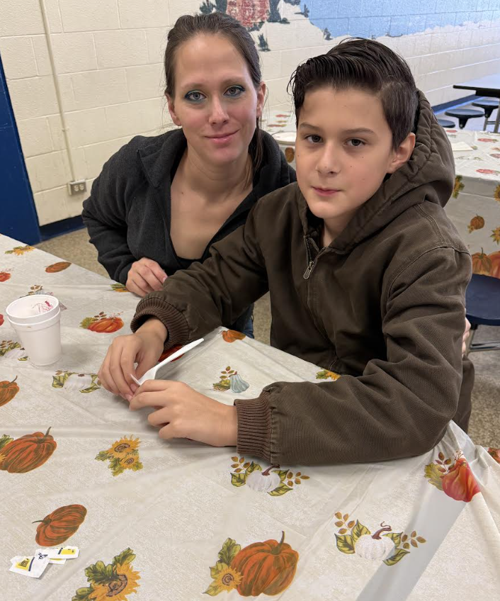 A woman and a boy sitting together at a table covered with a pumpkin-patterned tablecloth. The boy is wearing a brown hoodie, holding a pencil, and both are smiling softly toward the camera.